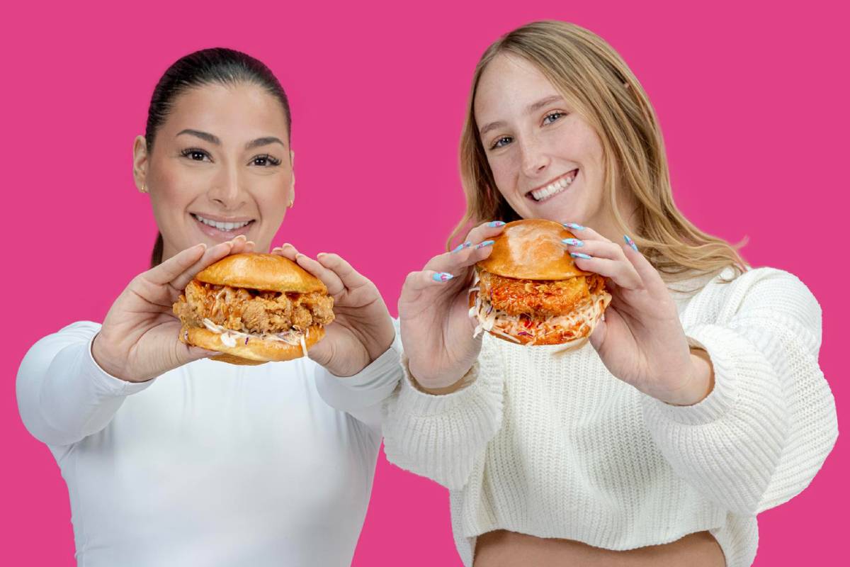 Two smiling women holding crispy fried chicken sandwiches on a pink background