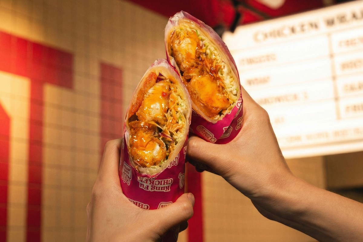 Two hands hold a chicken wrap cut in half, wrapped in pink paper, showing a hearty filling with crispy chicken, vegetables and sauce.